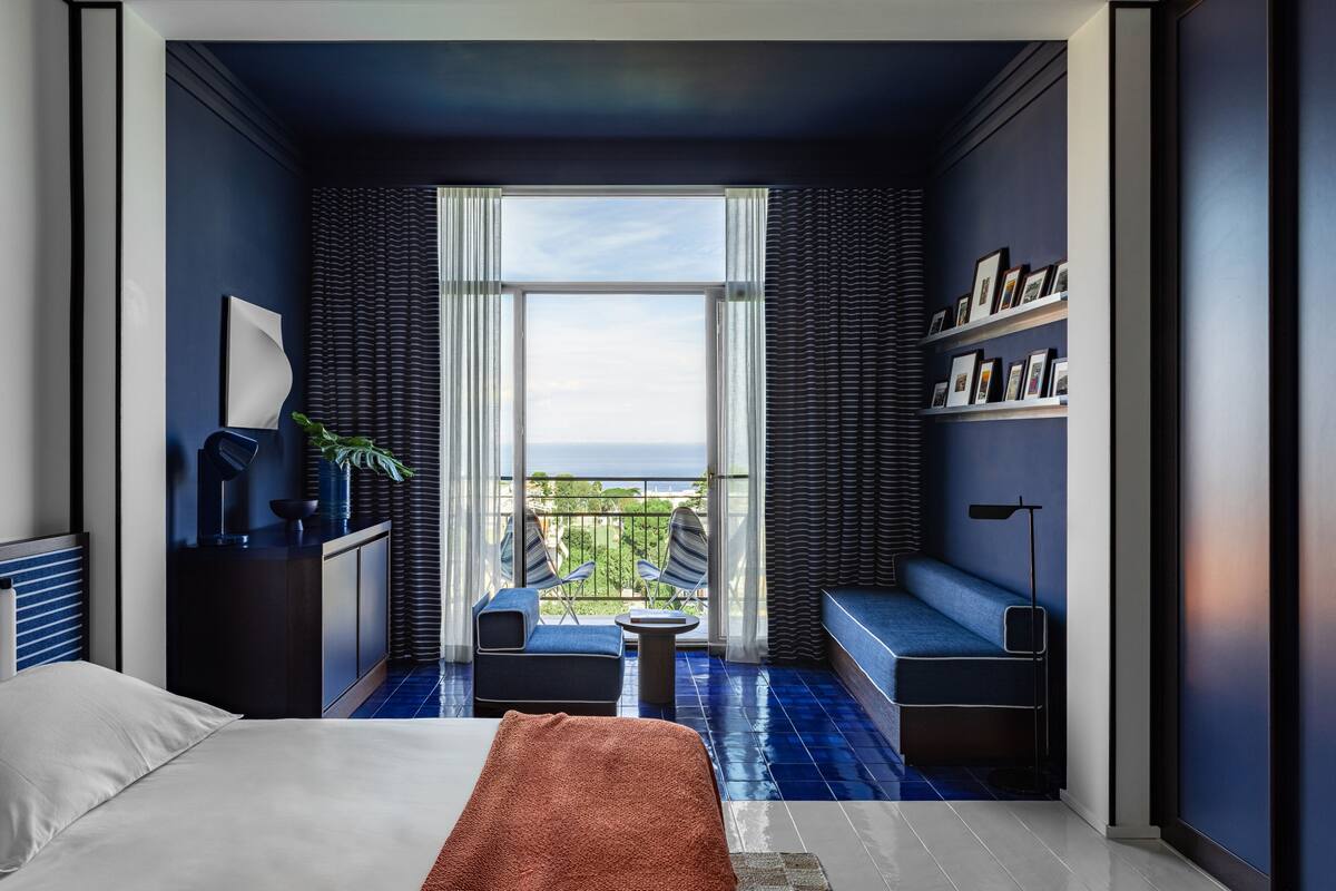 A modern hotel room with a large window, a bed, a seating area with a view, and a bookshelf.