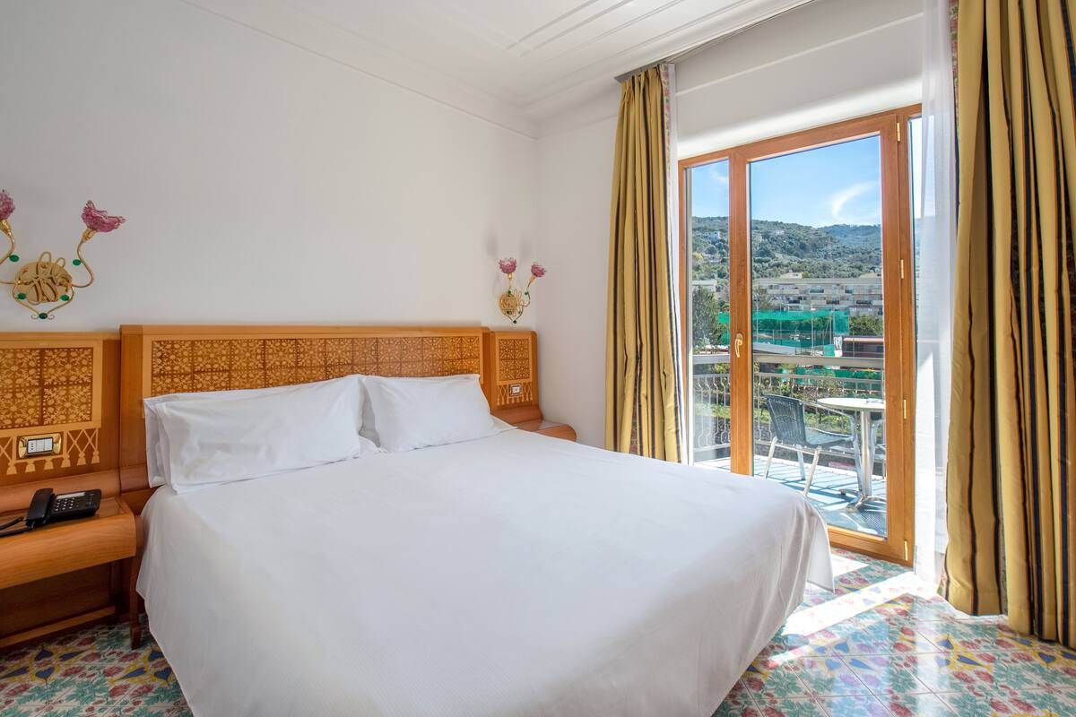 A hotel room with a large bed, a bedside phone, a decorative wall hanging, and a balcony with a view of the hills.