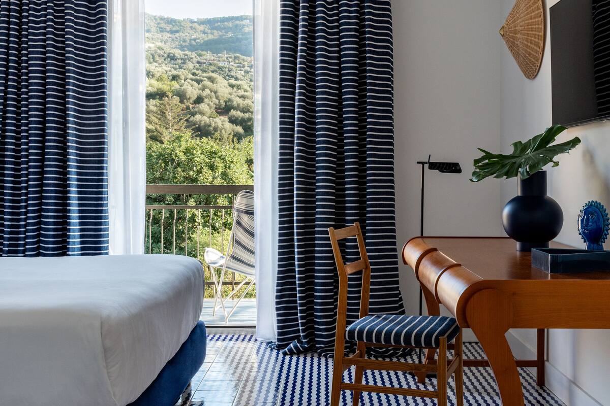 A hotel room with a bed, a wooden table, a chair, and a view of a mountainous landscape through the window.