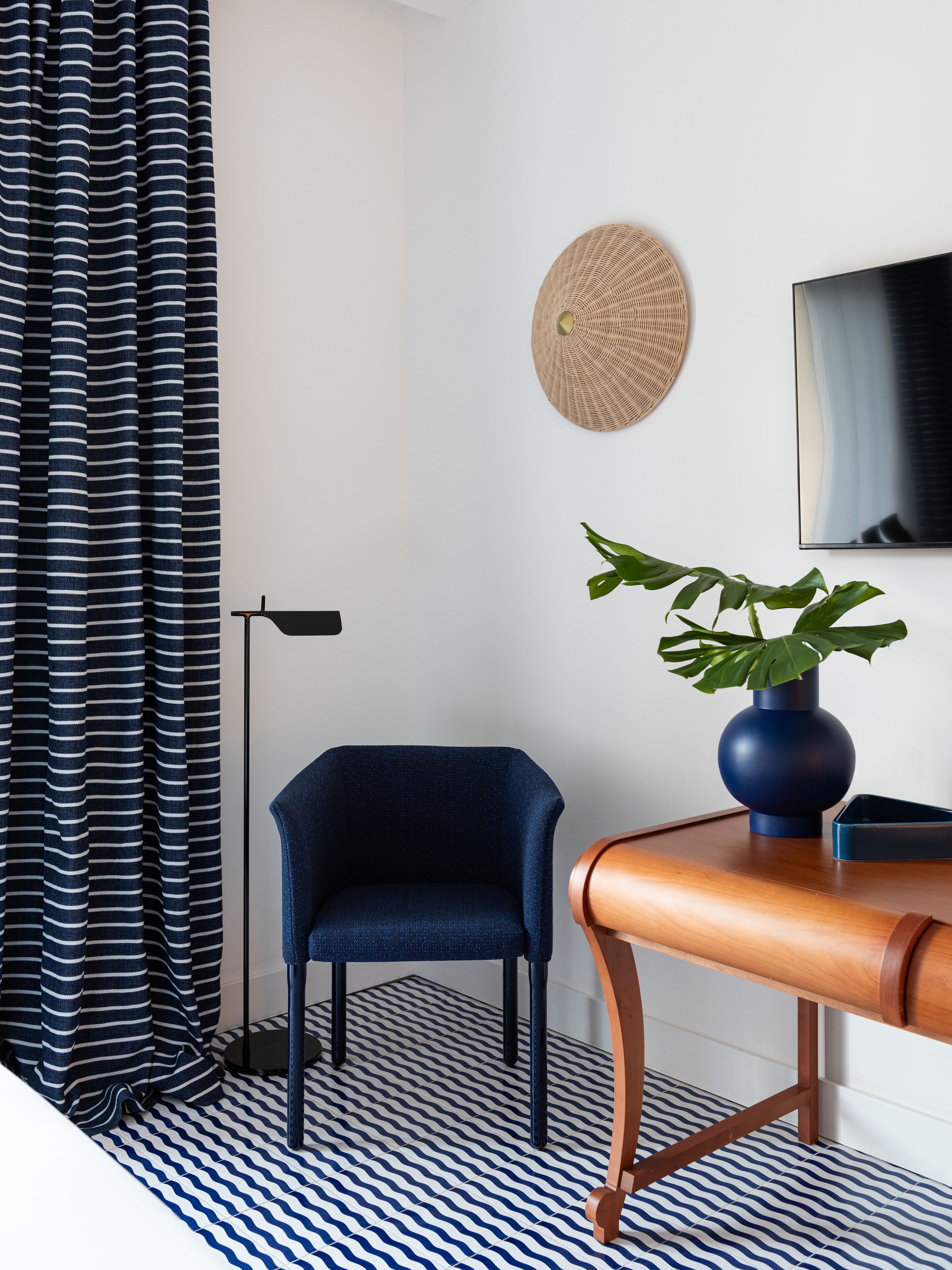 Una habitación moderna con cortinas a rayas, un adorno de pared redondo tejido, un sillón azul, una mesa de madera con un jarrón y un televisor en la pared.