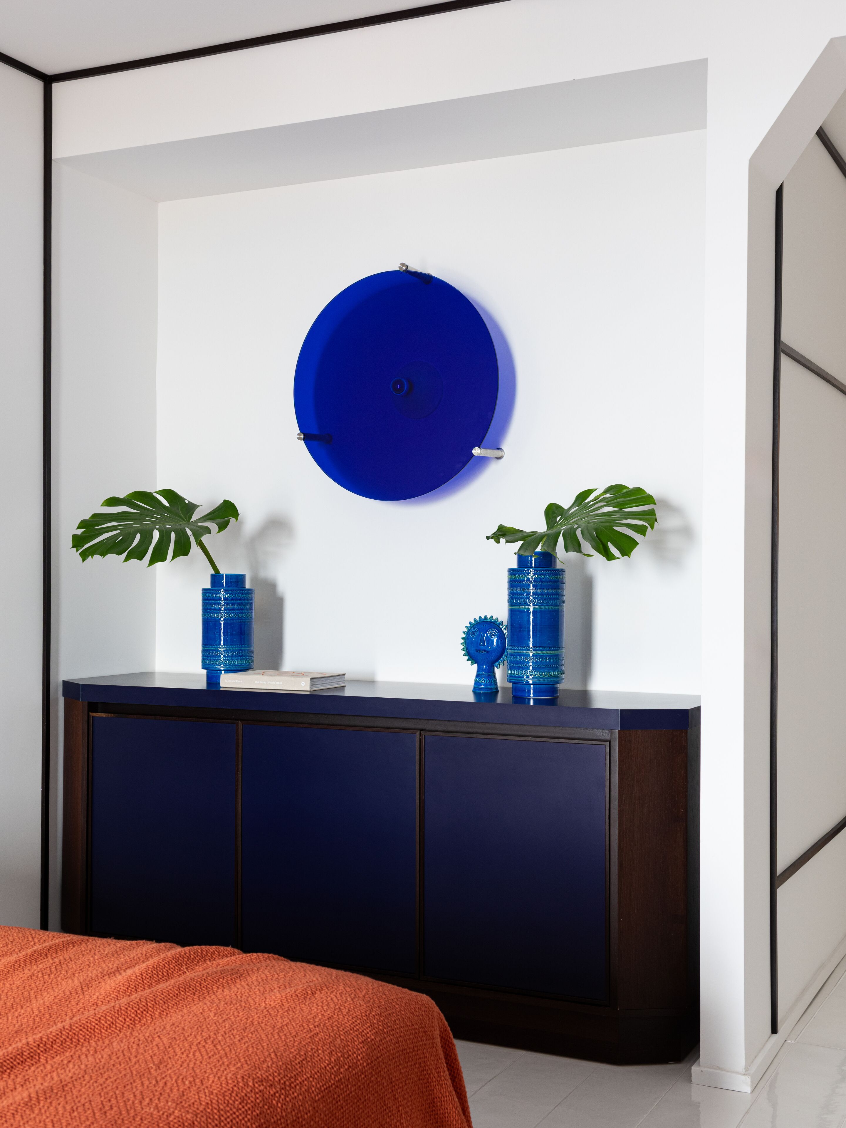 Una habitación moderna con una consola azul oscuro, un adorno circular azul en la pared y dos plantas verdes en macetas.