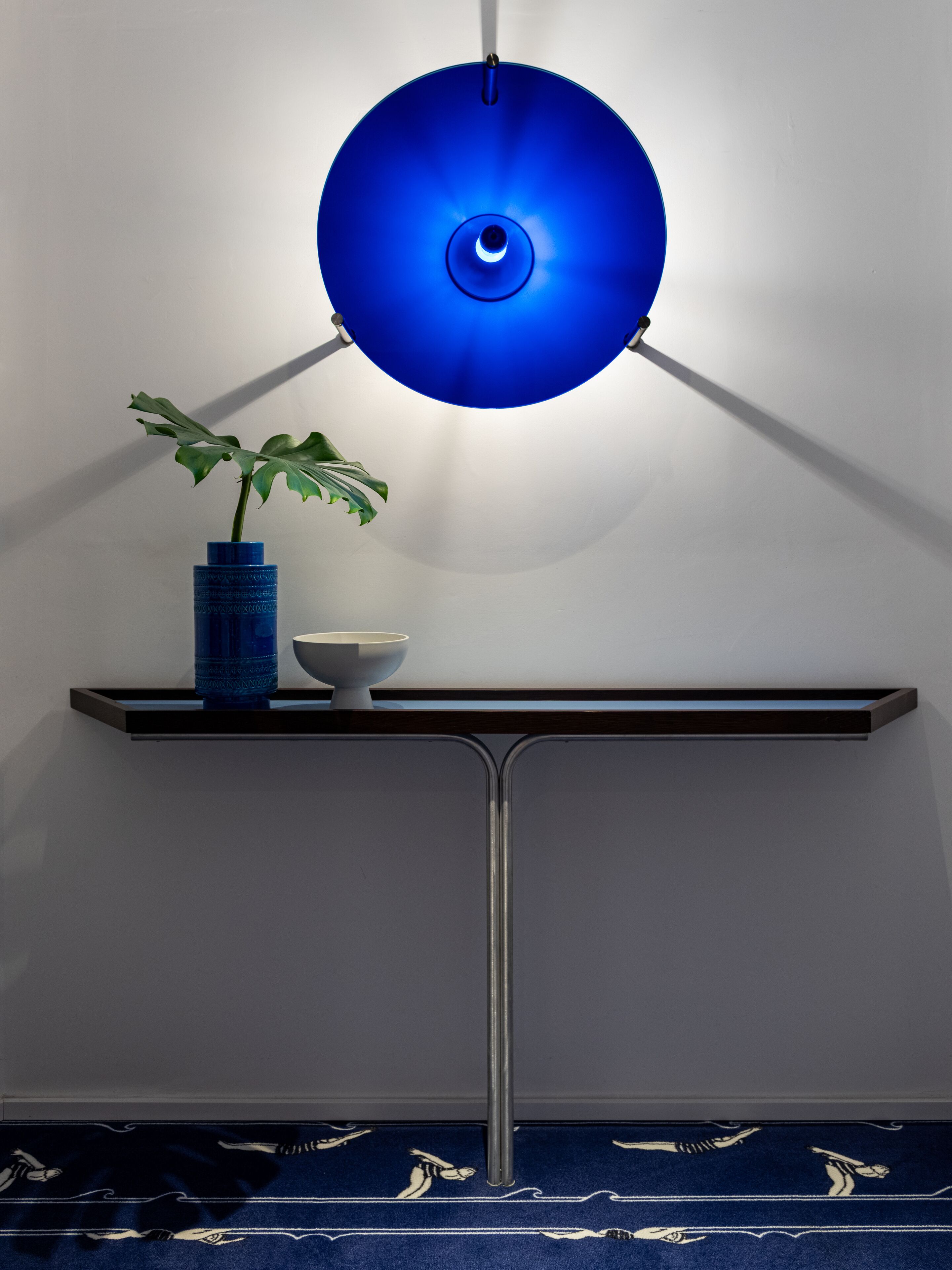 Ein modernes Zimmer mit einer blauen Pendelleuchte, einem Blumentopf und einer weißen Schale auf einem dunklen Konsolentisch.