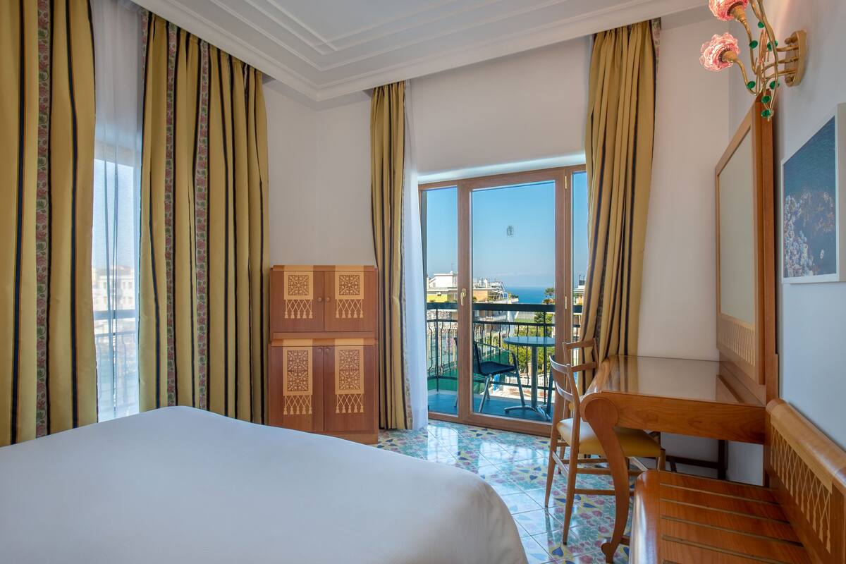 A hotel room with a bed, a wooden cabinet, a balcony with a table and chairs, and a view of the city.