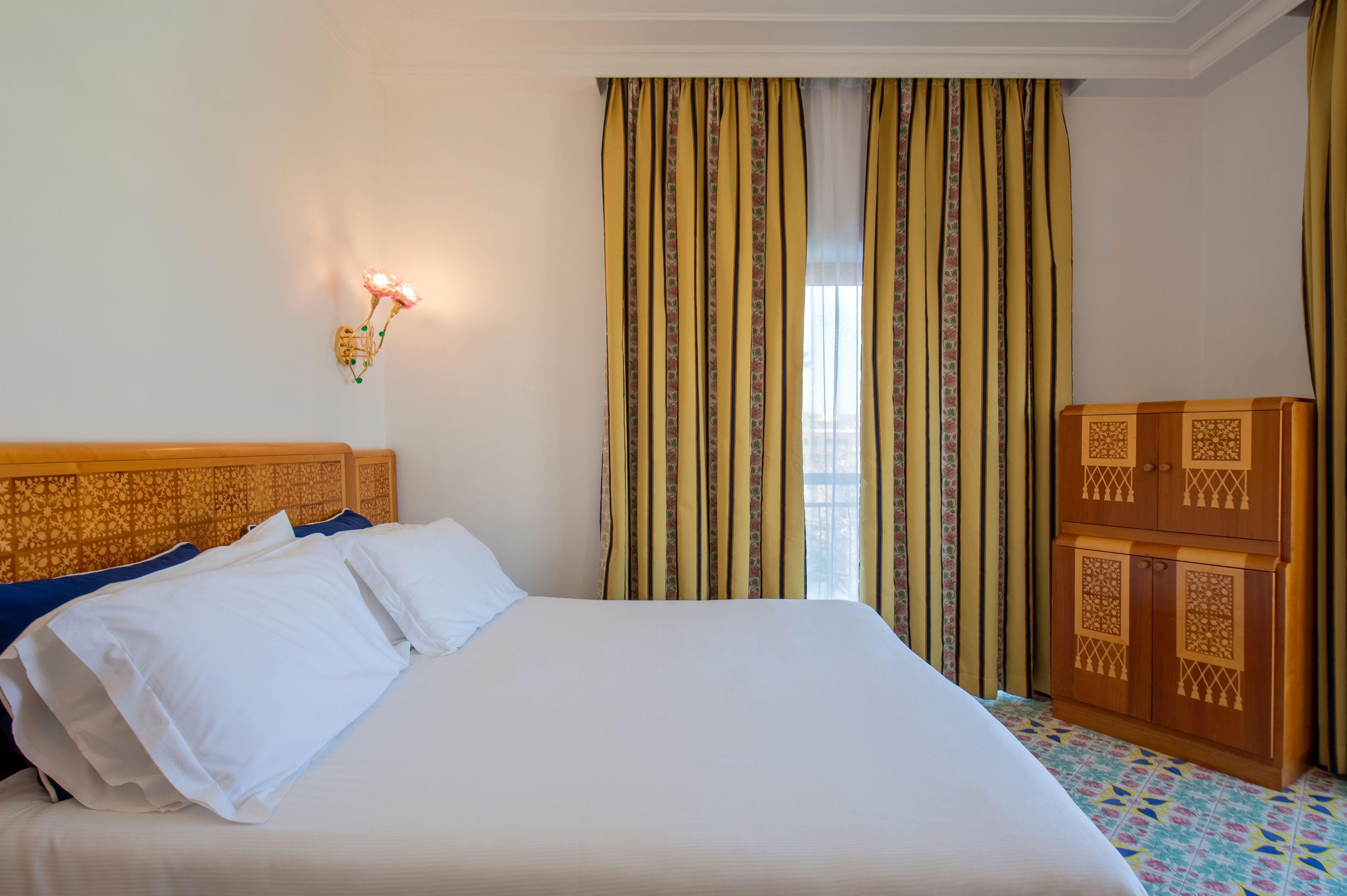 Quarto de hotel com uma cama grande, mesa de cabeceira, luminária na parede e janela com cortinas.