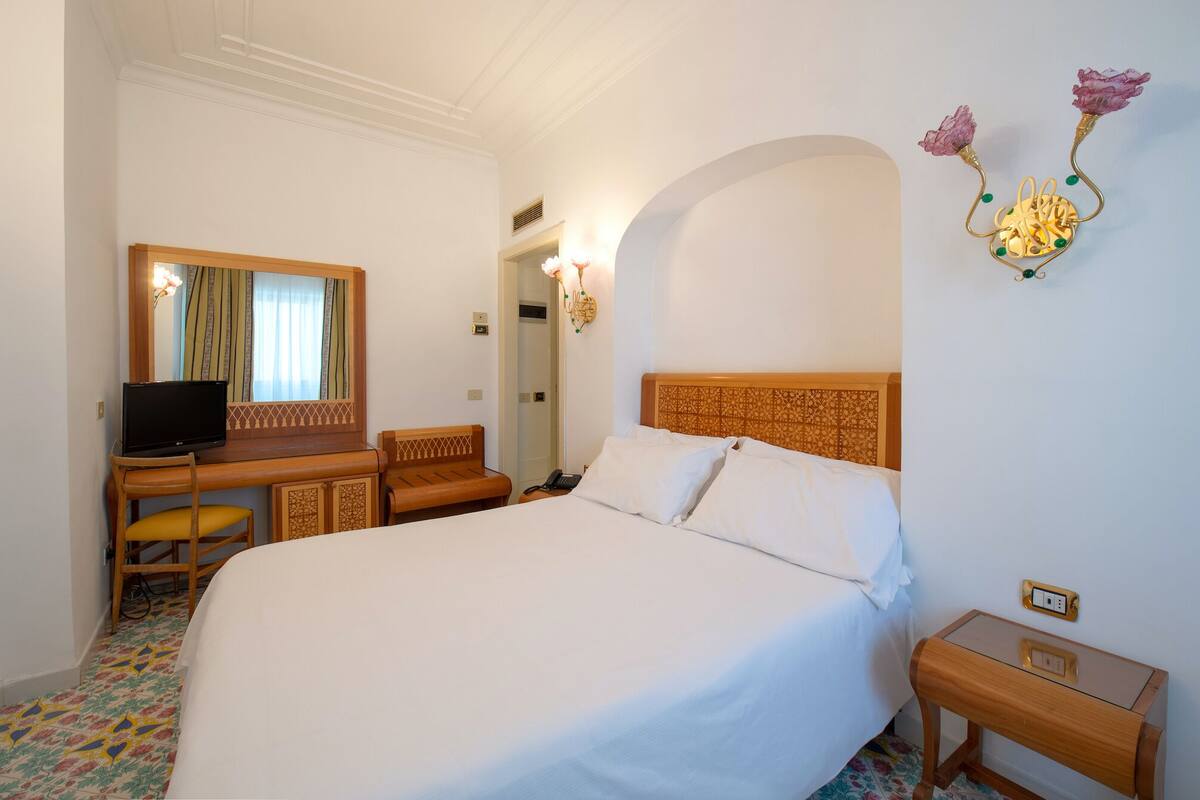 A hotel room with a large bed, a desk with a computer, a chair, a mirror, and a wall-mounted light fixture.
