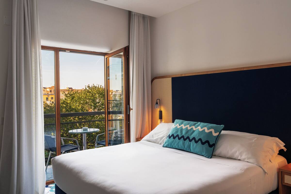 A hotel room with a large bed, a view of the outdoors through a sliding glass door, a small table, and a chair.