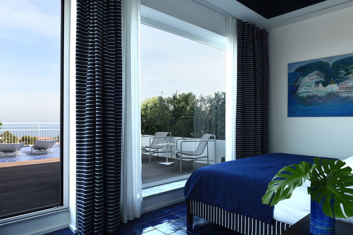 A bedroom with a large window, a balcony with outdoor furniture, and a painting on the wall.