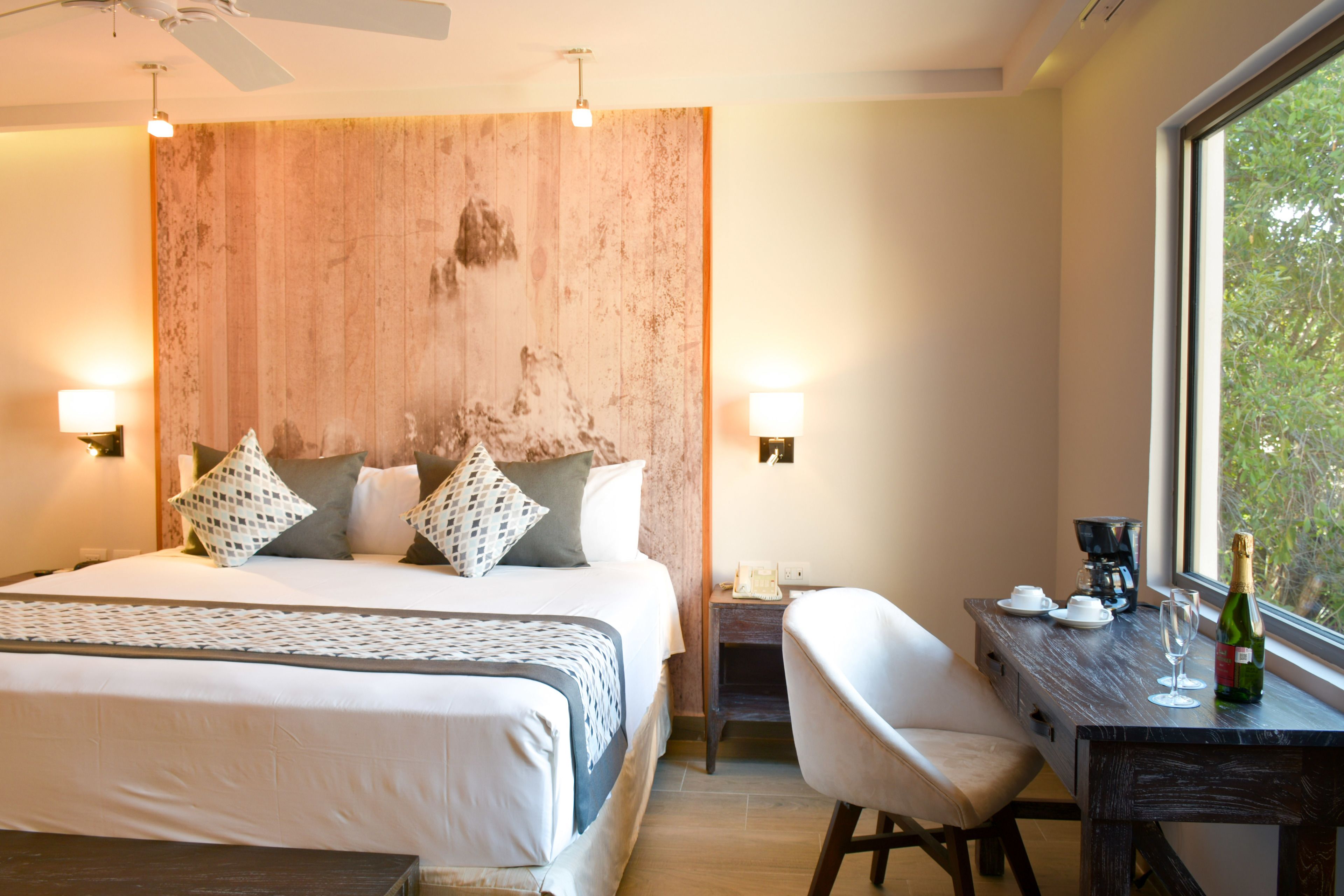 A hotel room with a large bed, bedside tables, a desk with a chair, a window with a view, and a ceiling fan.