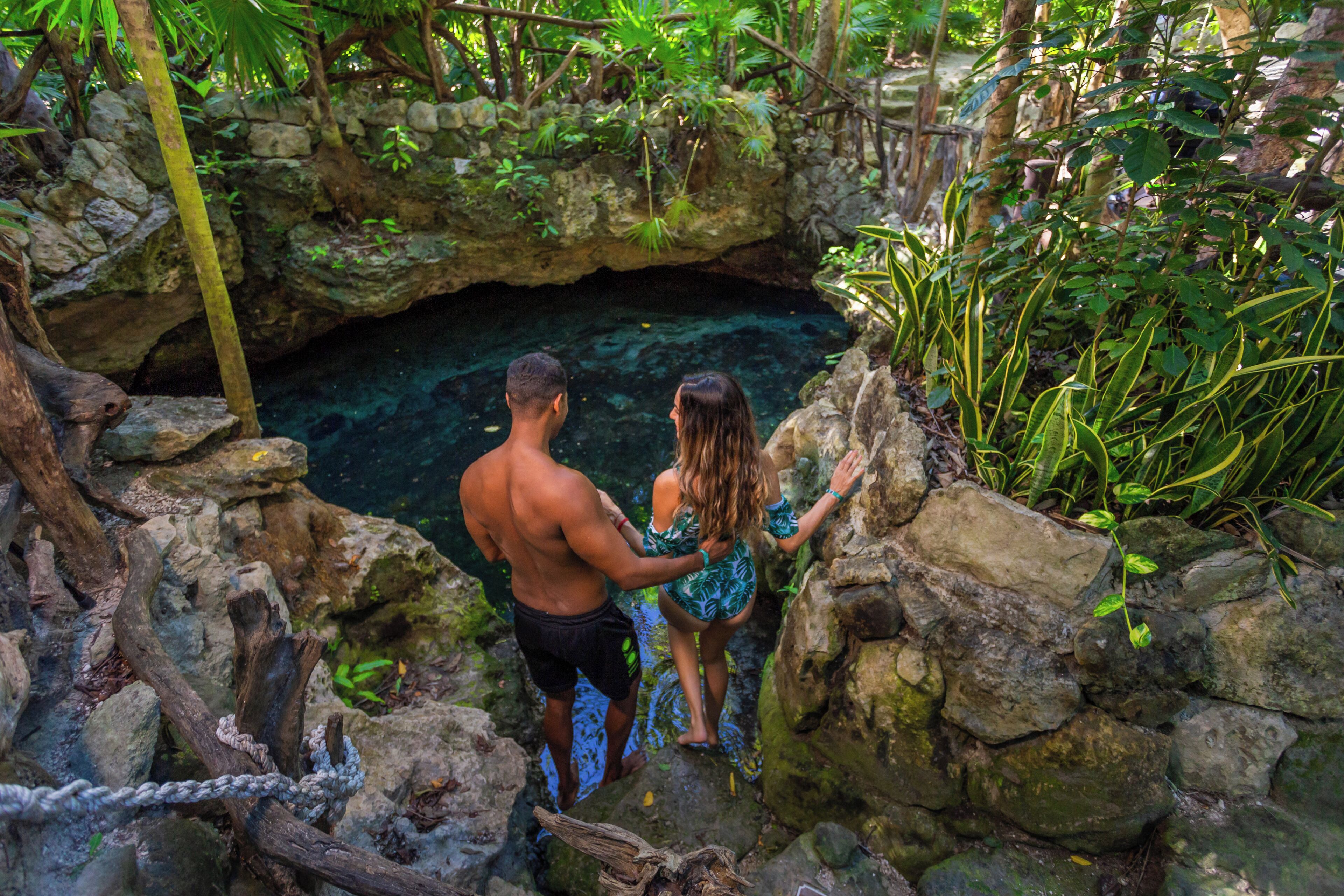 A man and a woman standing at the edge of a rocky cave with a clear water pool inside.