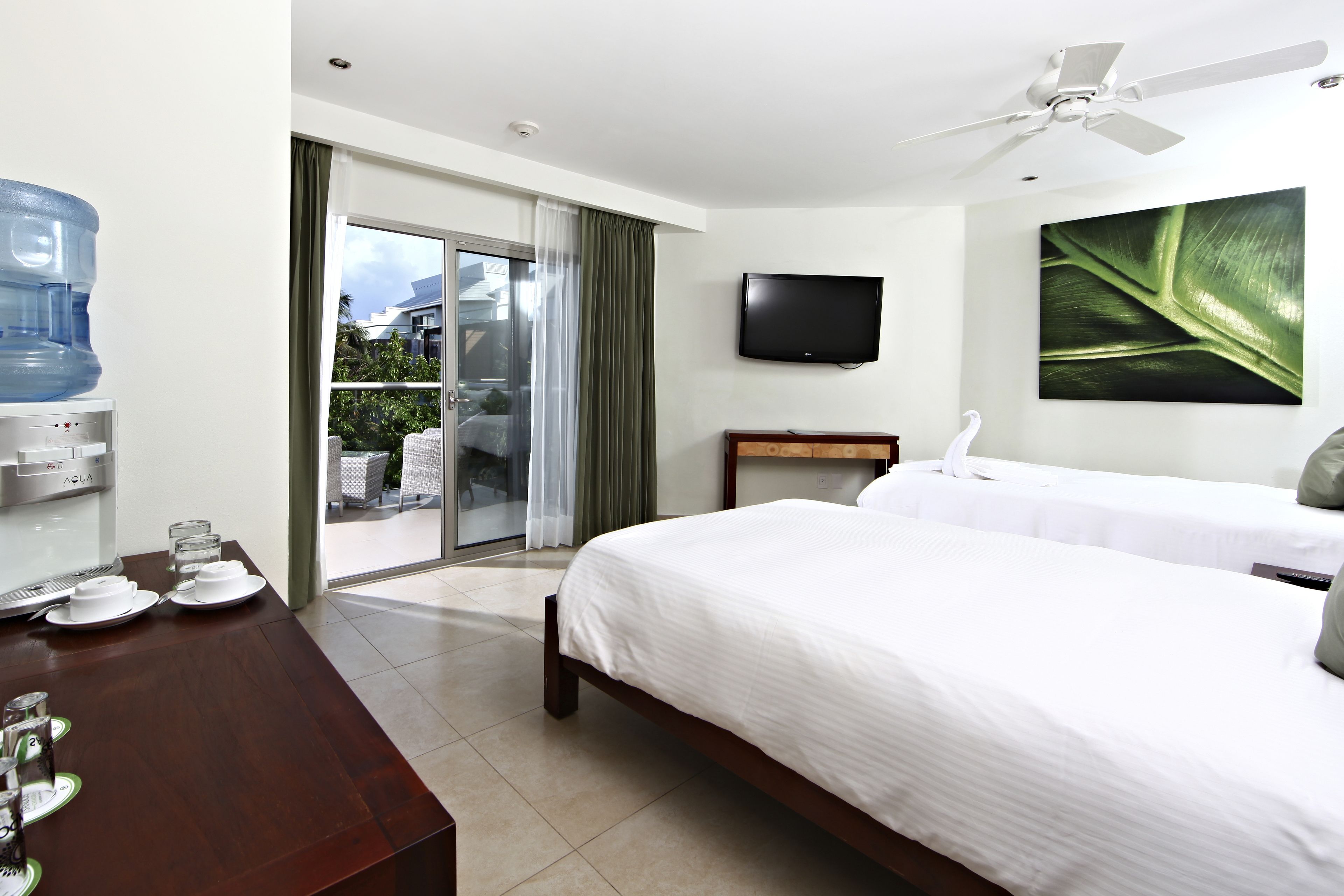 A hotel room with a bed, a desk with a water dispenser, a flat-screen TV, and a large window with a view of the outside.