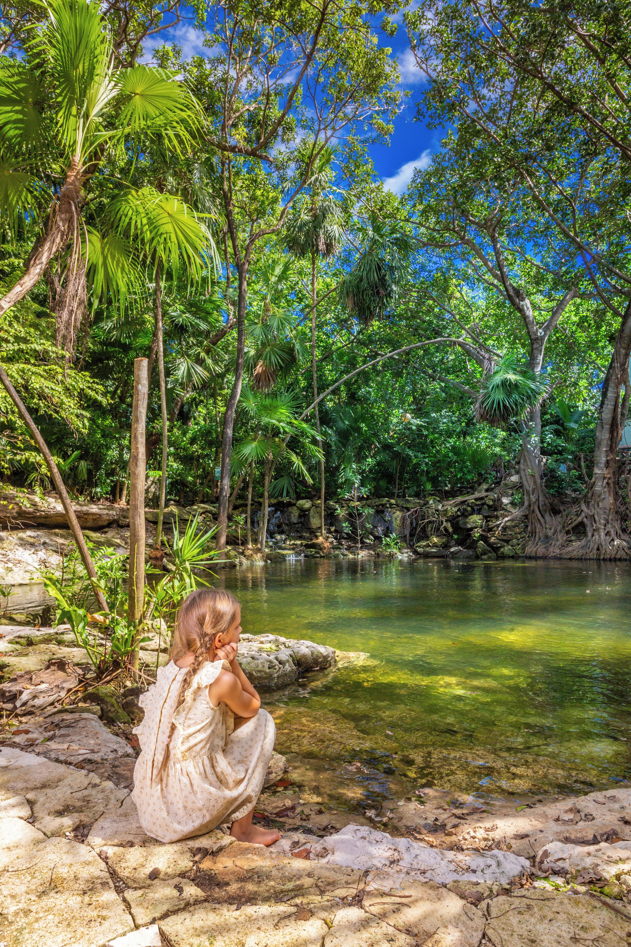 A natural setting with a clear water body, trees, and a person sitting by the water.