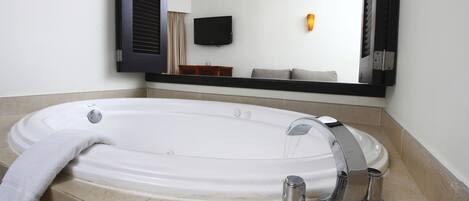 A modern bathroom with a large bathtub, a mirror, and a flat-screen TV mounted on the wall.