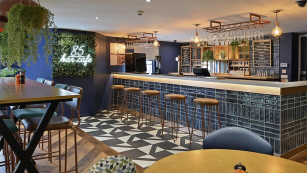 A bar area with a counter, stools, and a menu board listing drinks. - Travelodge Leeds Vicar Lane (Leeds)