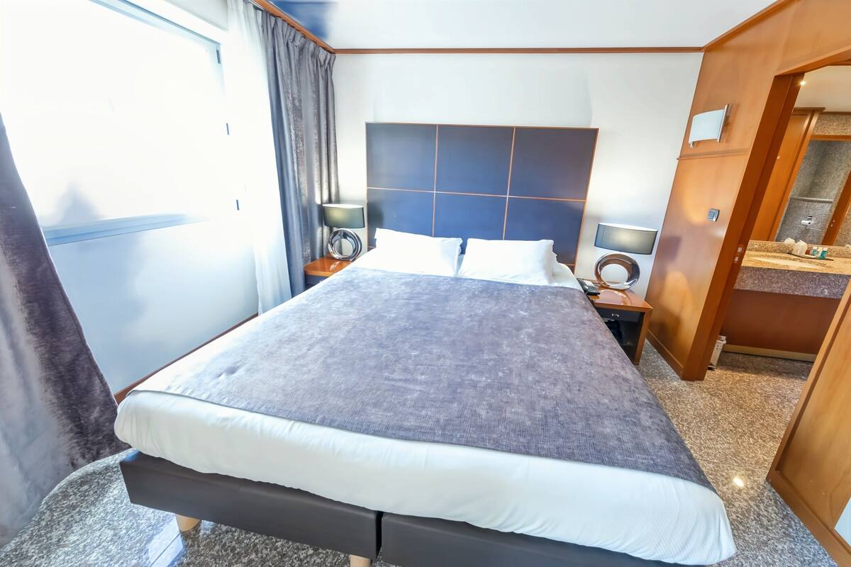 A hotel room with a large bed, a bedside table with a lamp, a small desk with a mirror, and a bathroom visible through an open door.