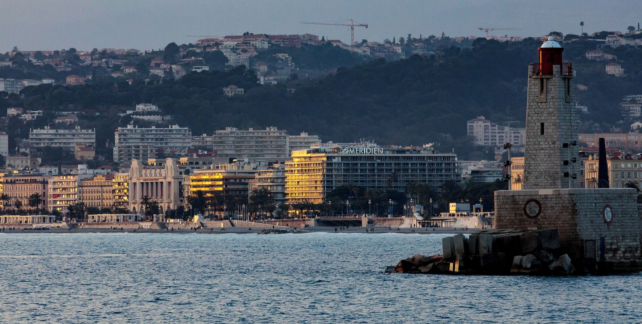 Un paisaje urbano costero con un faro destacado, un hotel llamado 'LEMERIDIEN' y varios edificios a lo largo del paseo marítimo.