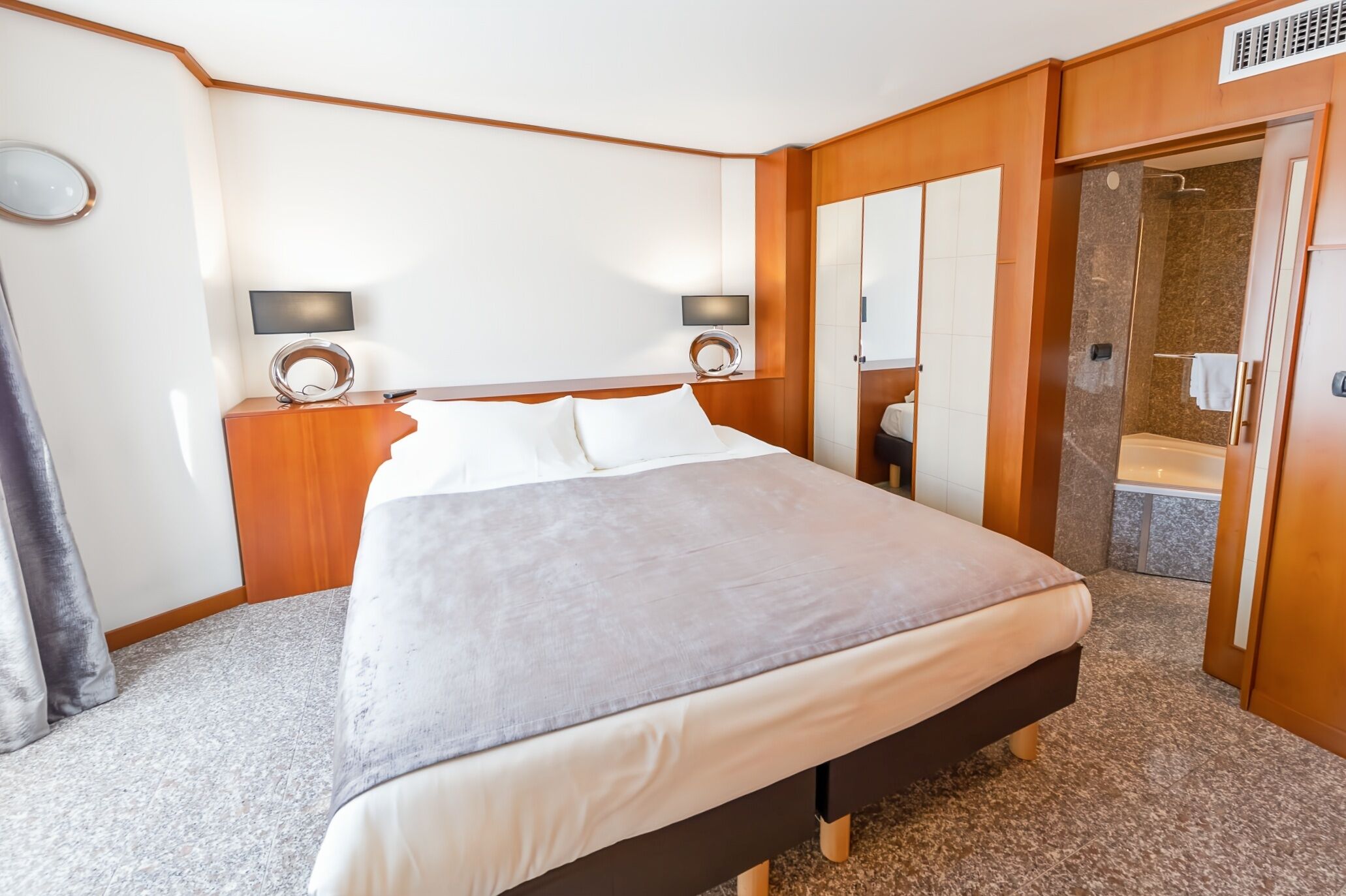Une chambre d’hôtel avec un grand lit, deux tables de chevet avec des lampes, un miroir et une salle de bain visible à travers une porte ouverte.