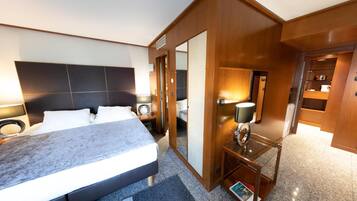 A modern hotel room with a large bed, a bedside table with a lamp, a mirror, and a small seating area with a book.