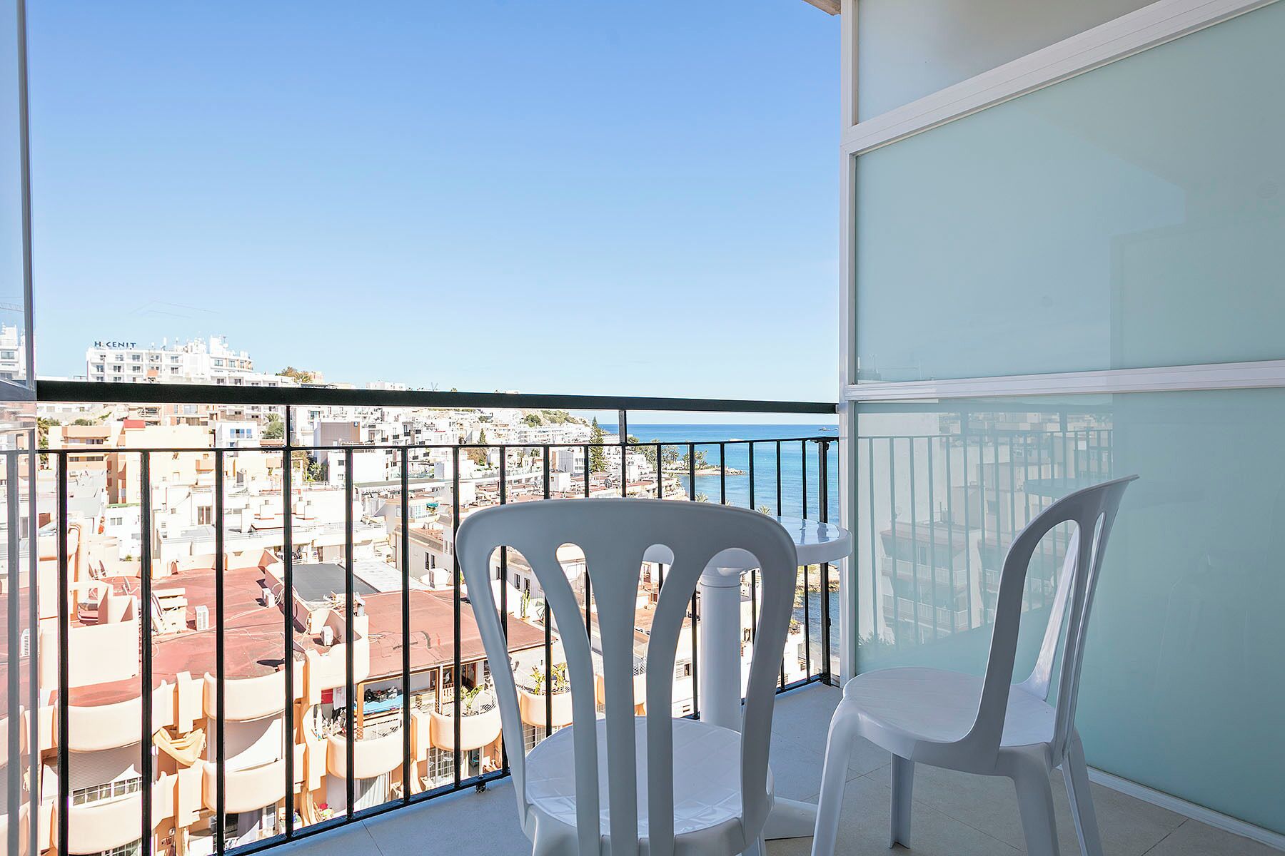 Un balcon avec deux chaises blanches, une balustrade en verre et une vue sur le paysage urbain et la mer.