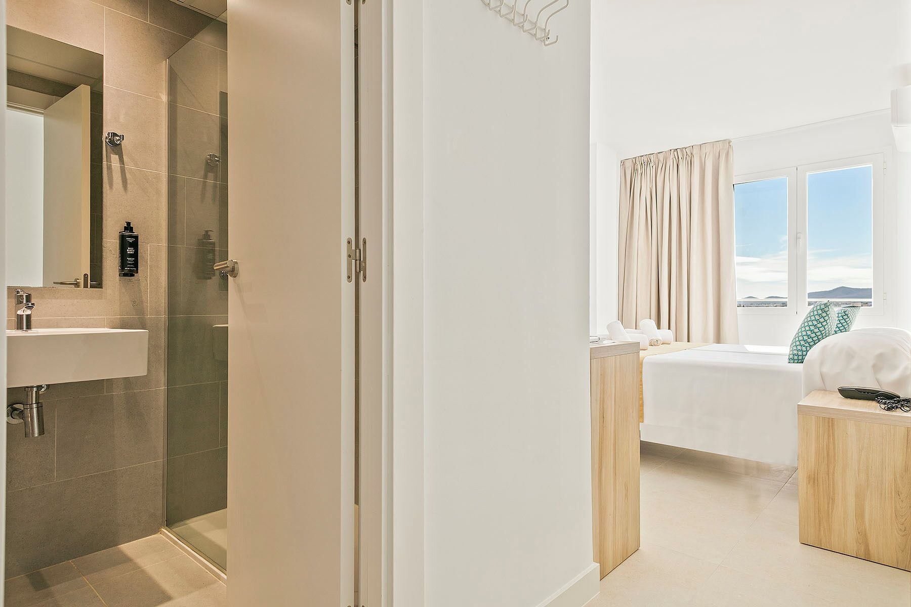 A hotel room with a bathroom, a bed, a bedside table, a window with curtains, and a view.