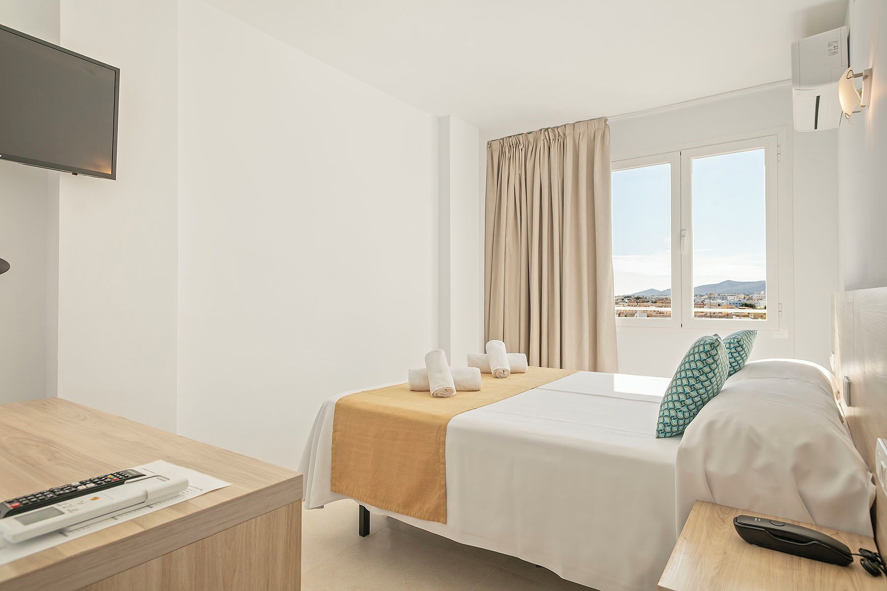 A hotel room with a bed, a desk, a TV, a window with curtains, and a view of the city.