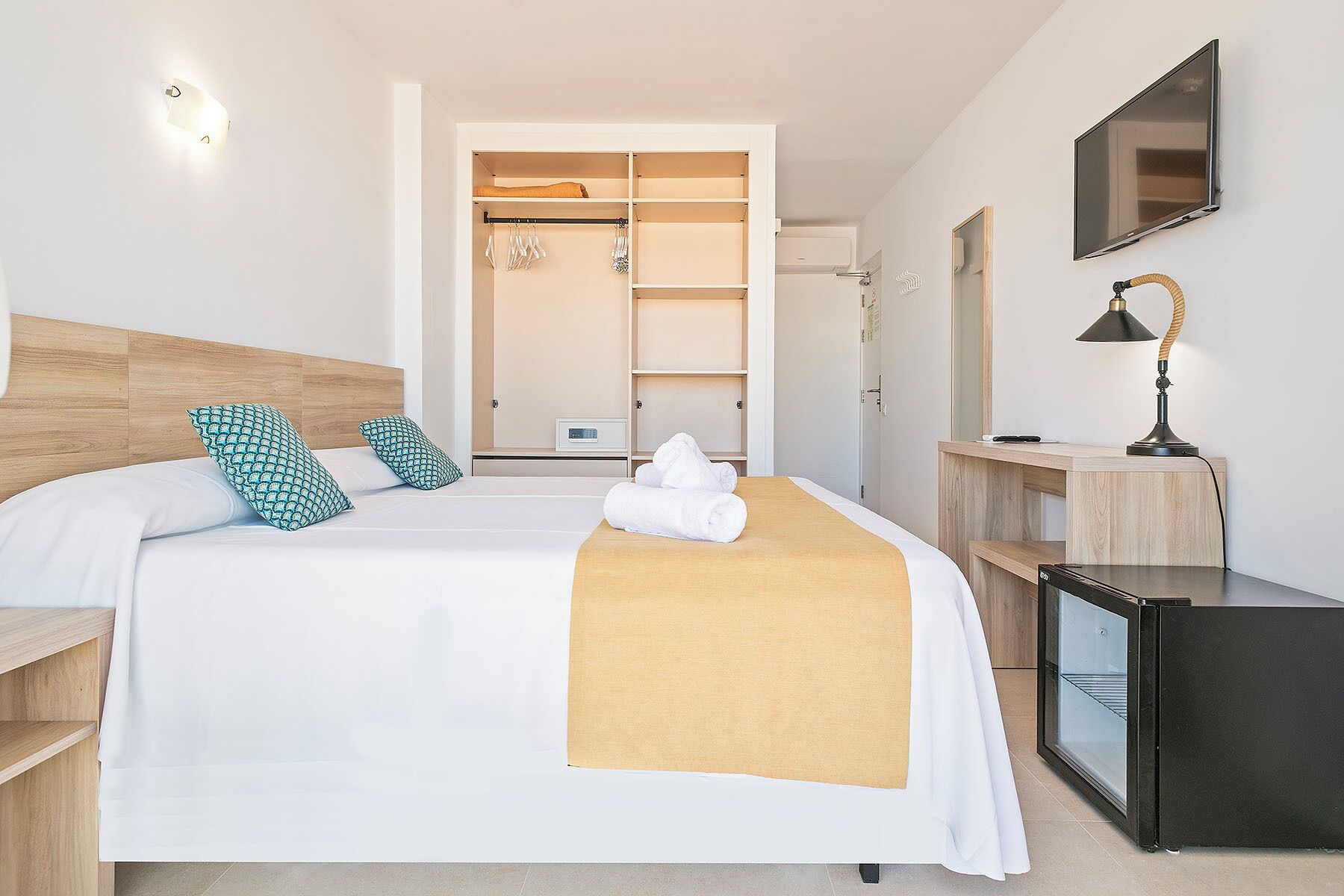 A hotel room with a bed, a desk with a lamp, a TV, and a minibar.