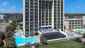 A multi-story hotel with a rooftop pool area, including lounge chairs and a glass-enclosed section. - Ocean Park Resort by Vacasa (Myrtle Beach)