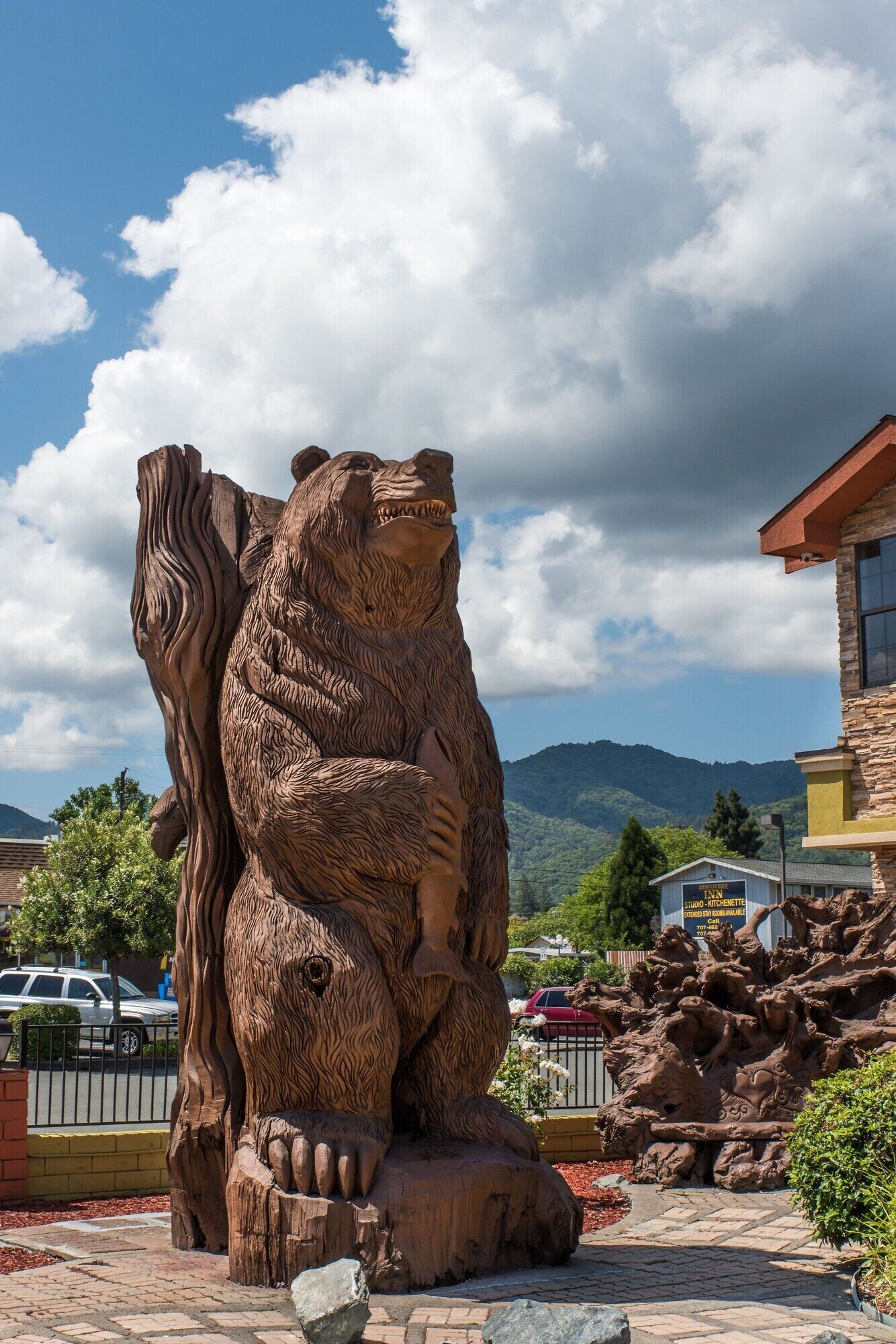 A large wooden sculpture of a bear holding a child, located in a landscaped area with a mountain backdrop.