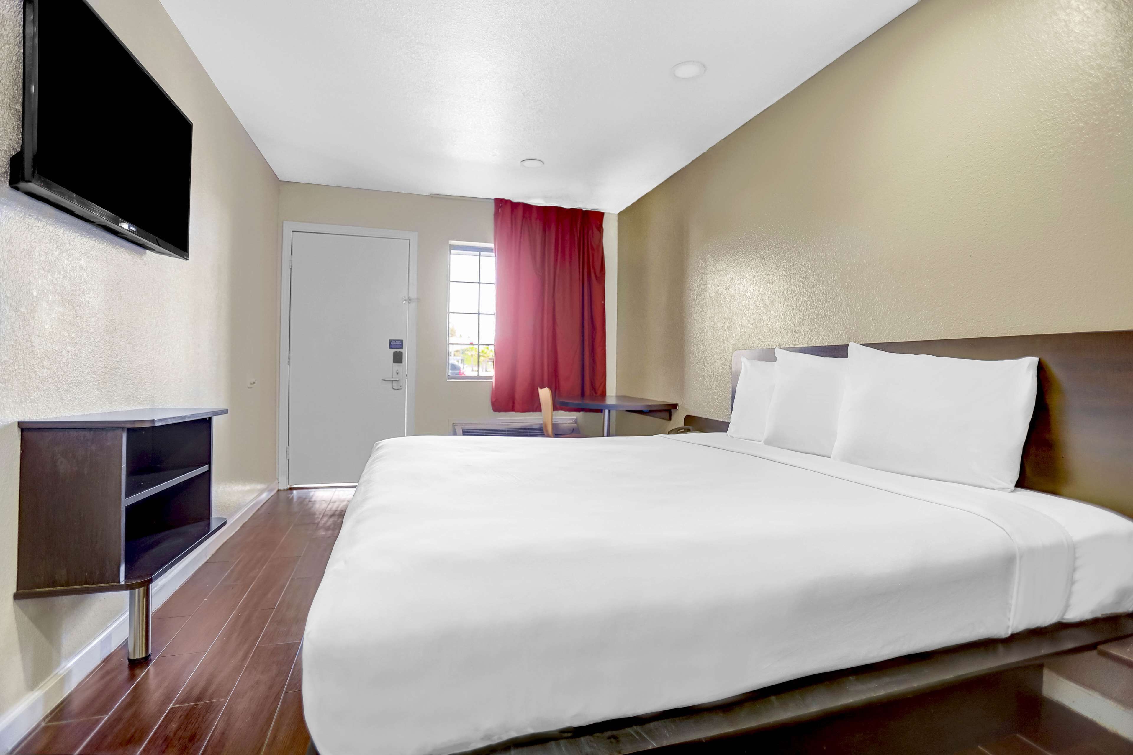 A hotel room with a large bed, a TV mounted on the wall, a wooden nightstand, and a window with red curtains.