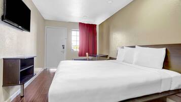 A hotel room with a large bed, a TV mounted on the wall, a wooden nightstand, and a window with red curtains.
