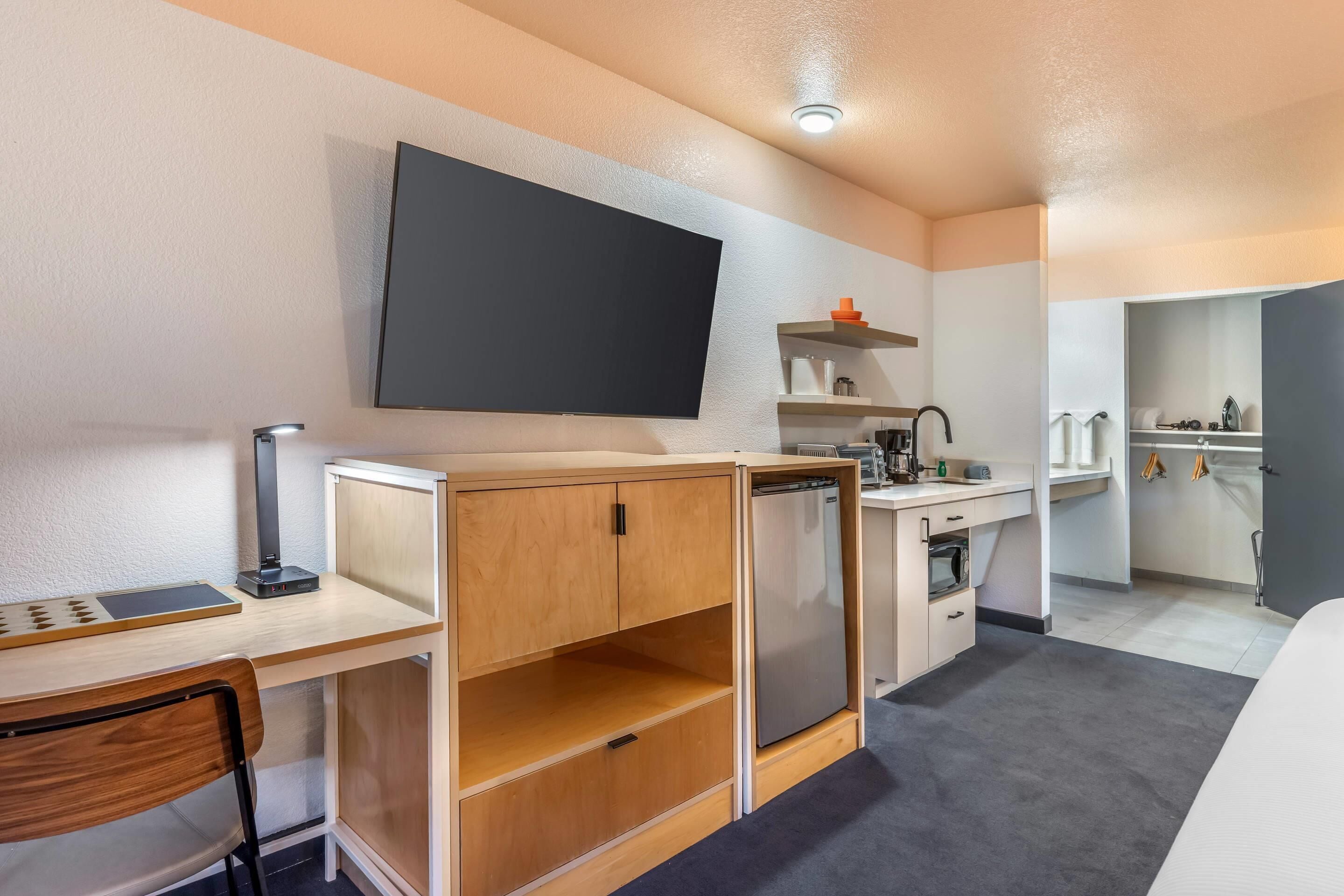 A compact hotel room with a desk, chair, TV, kitchenette, and a bed.