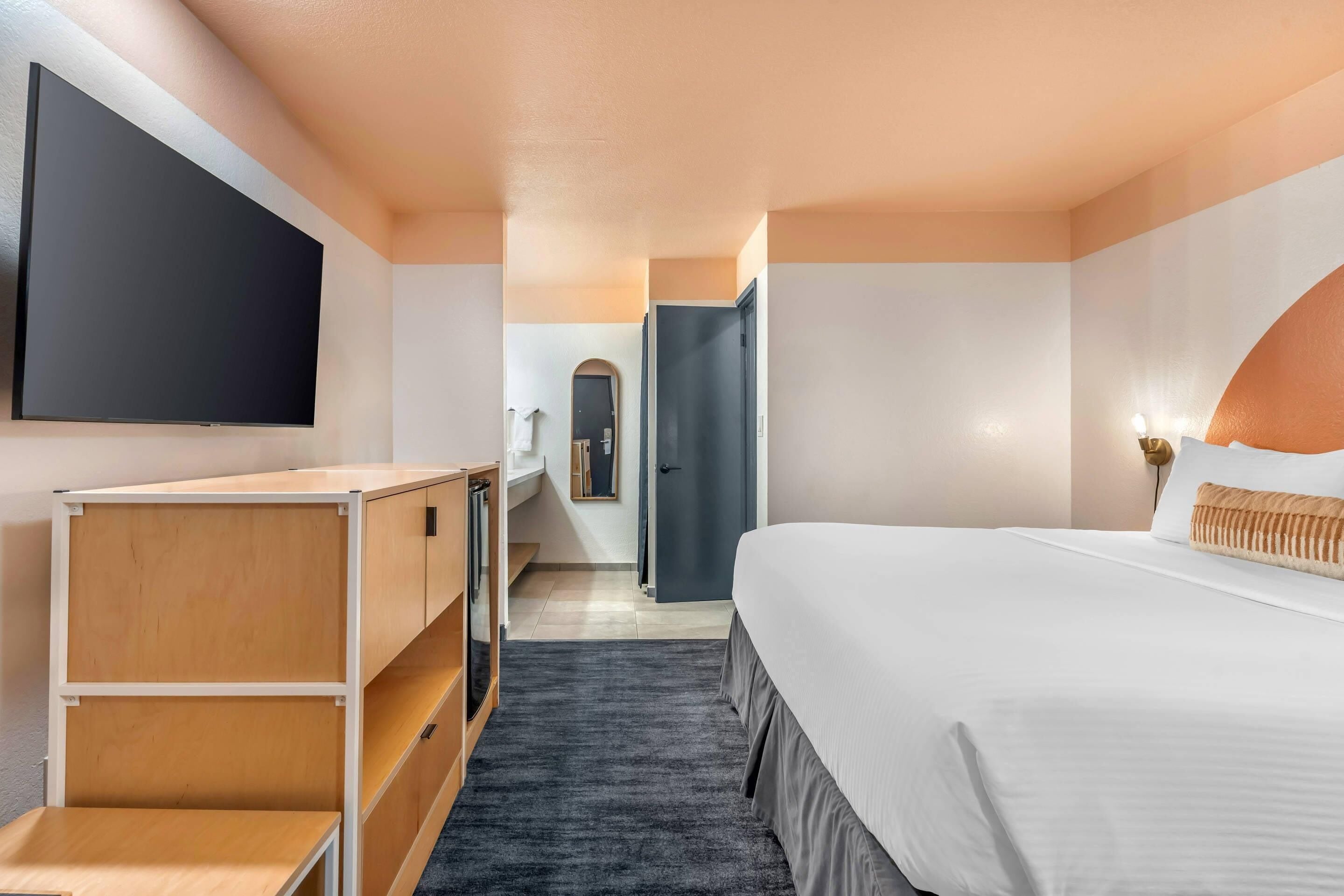 A modern hotel room with a large bed, a flat-screen TV mounted on the wall, a wooden desk, and a walk-in closet.