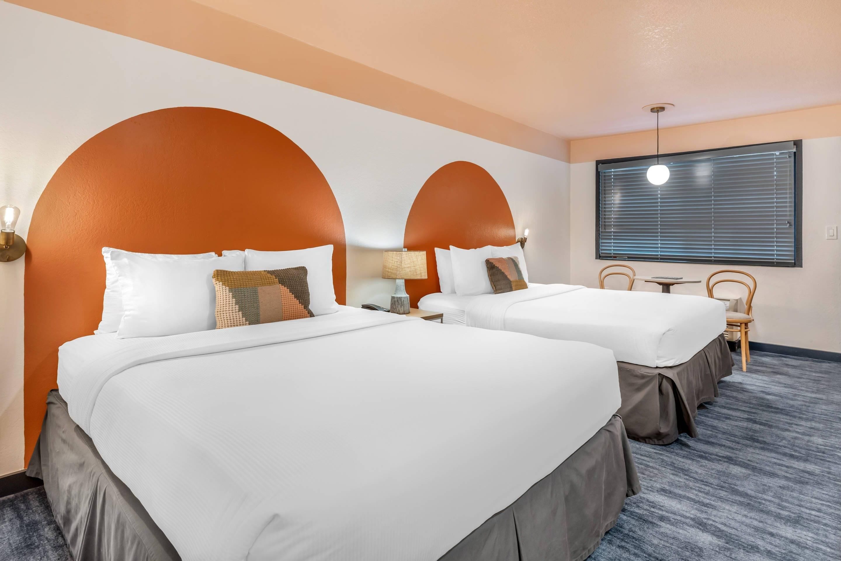 A hotel room with two beds, a small table, and a window with blinds.