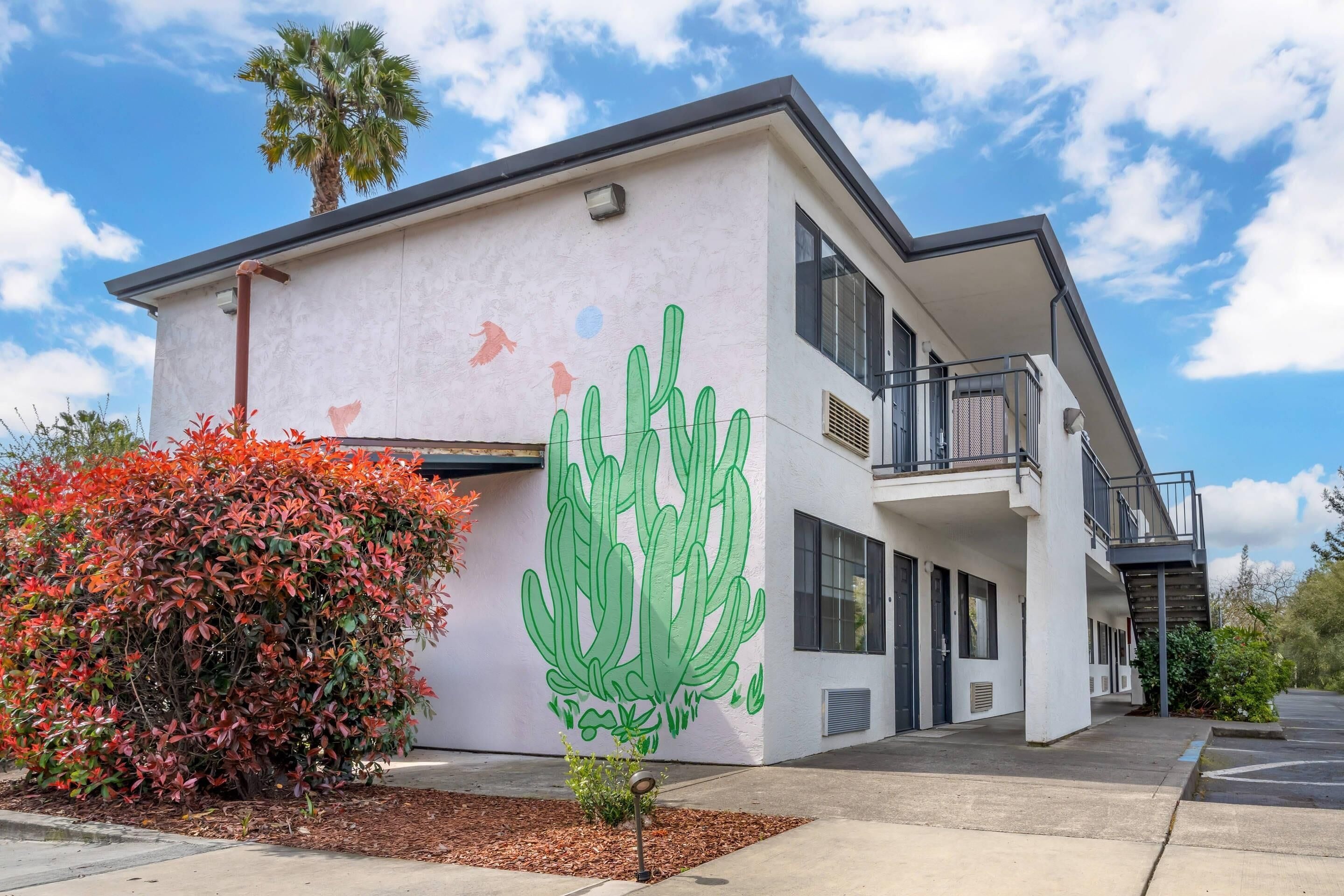 A modern building with a white exterior, a large green cactus mural, and a red flowering bush.