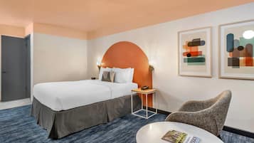 Een hotelkamer met een bed, een ronde tafel met een tijdschrift, een stoel en twee ingelijste abstracte kunstwerken aan de muur.