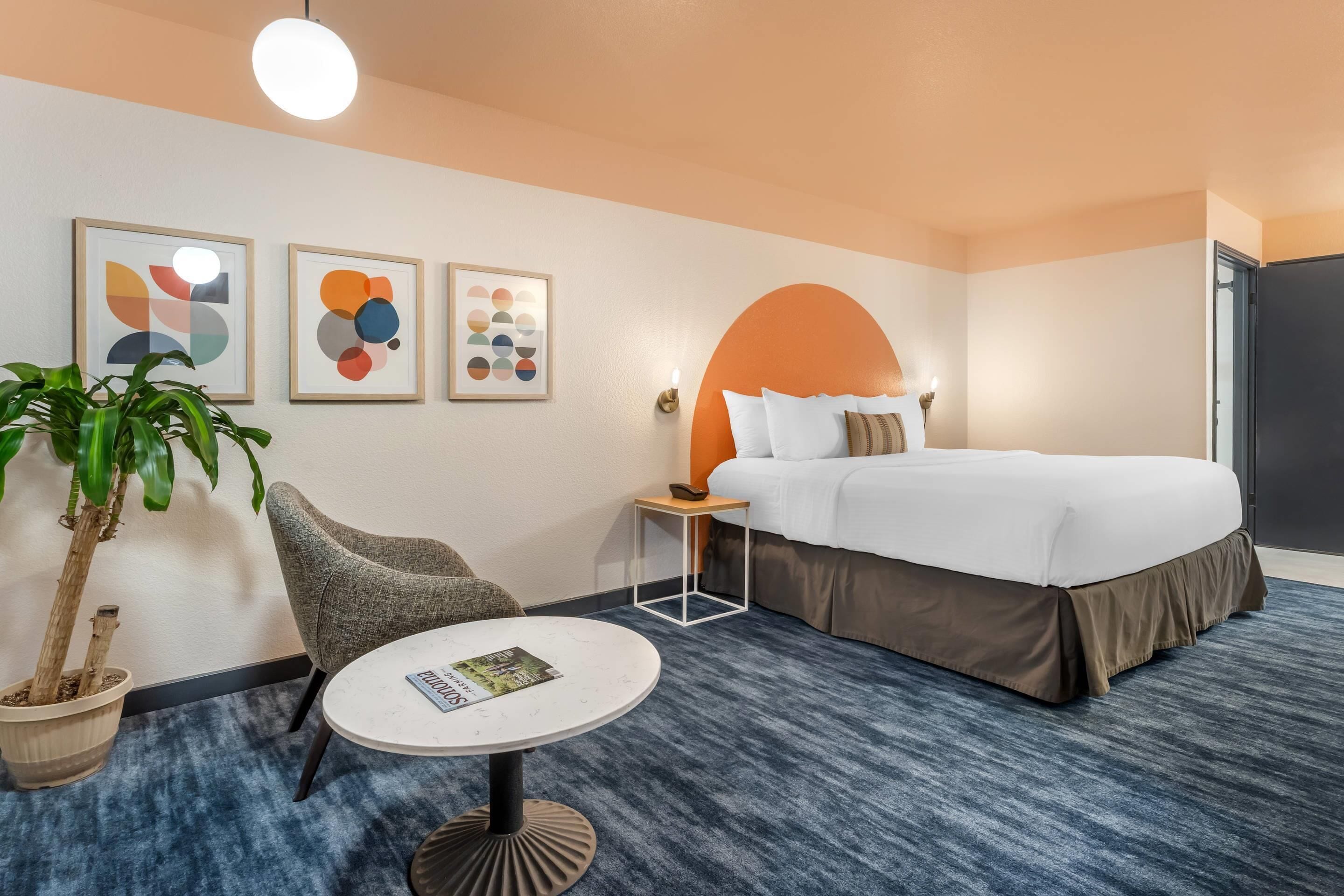 A hotel room with a bed, a round table with a magazine, a potted plant, a grey armchair, and abstract wall art.