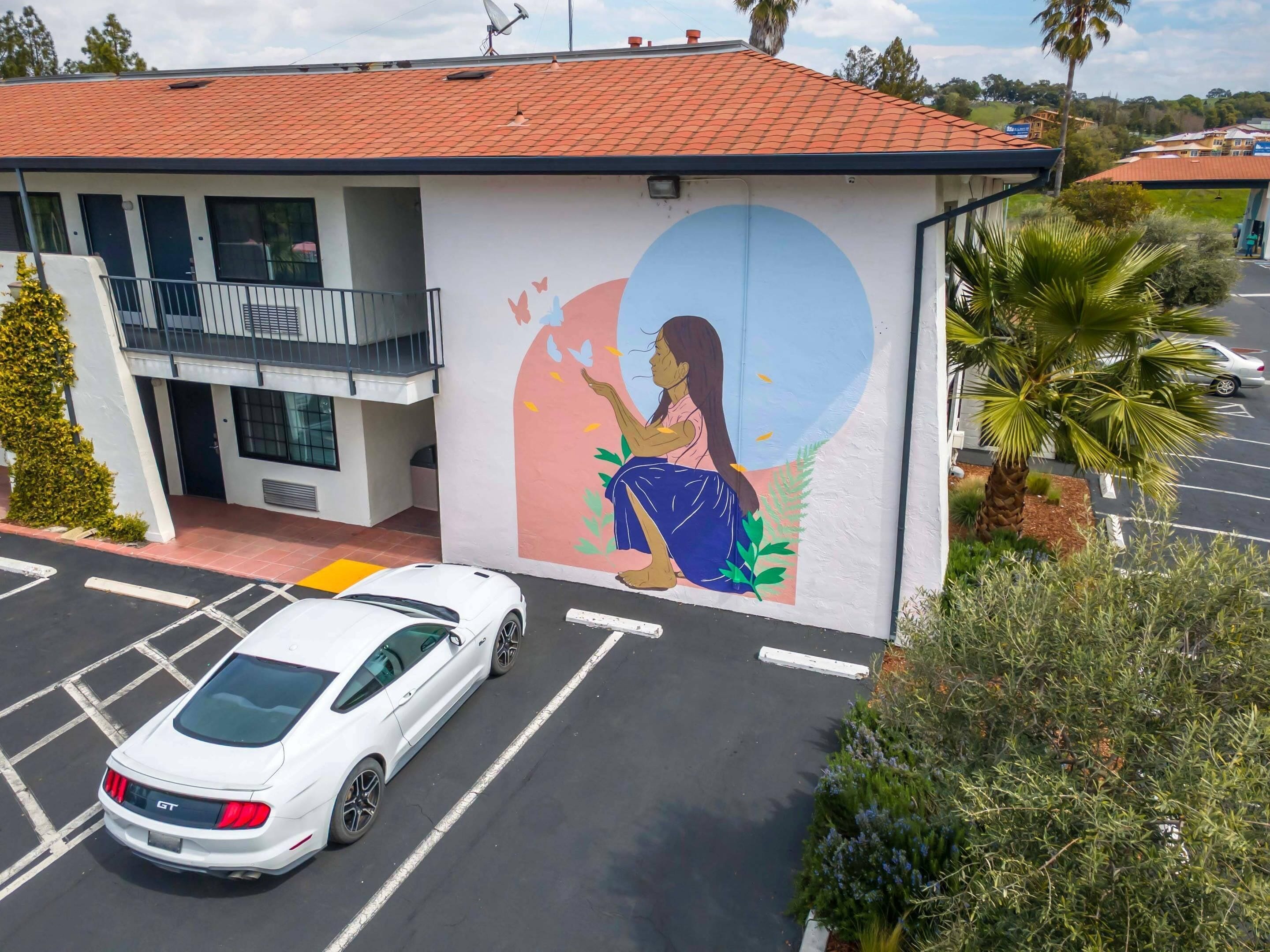 A building with a mural of a woman sitting on a bench, a parking lot, and a white car parked in front.