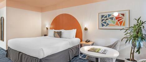 A hotel room with a large bed, a small table with a magazine, a potted plant, and a wall art piece.