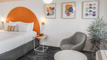 A modern hotel room with a large orange headboard, a grey armchair, a small table, and a potted plant.