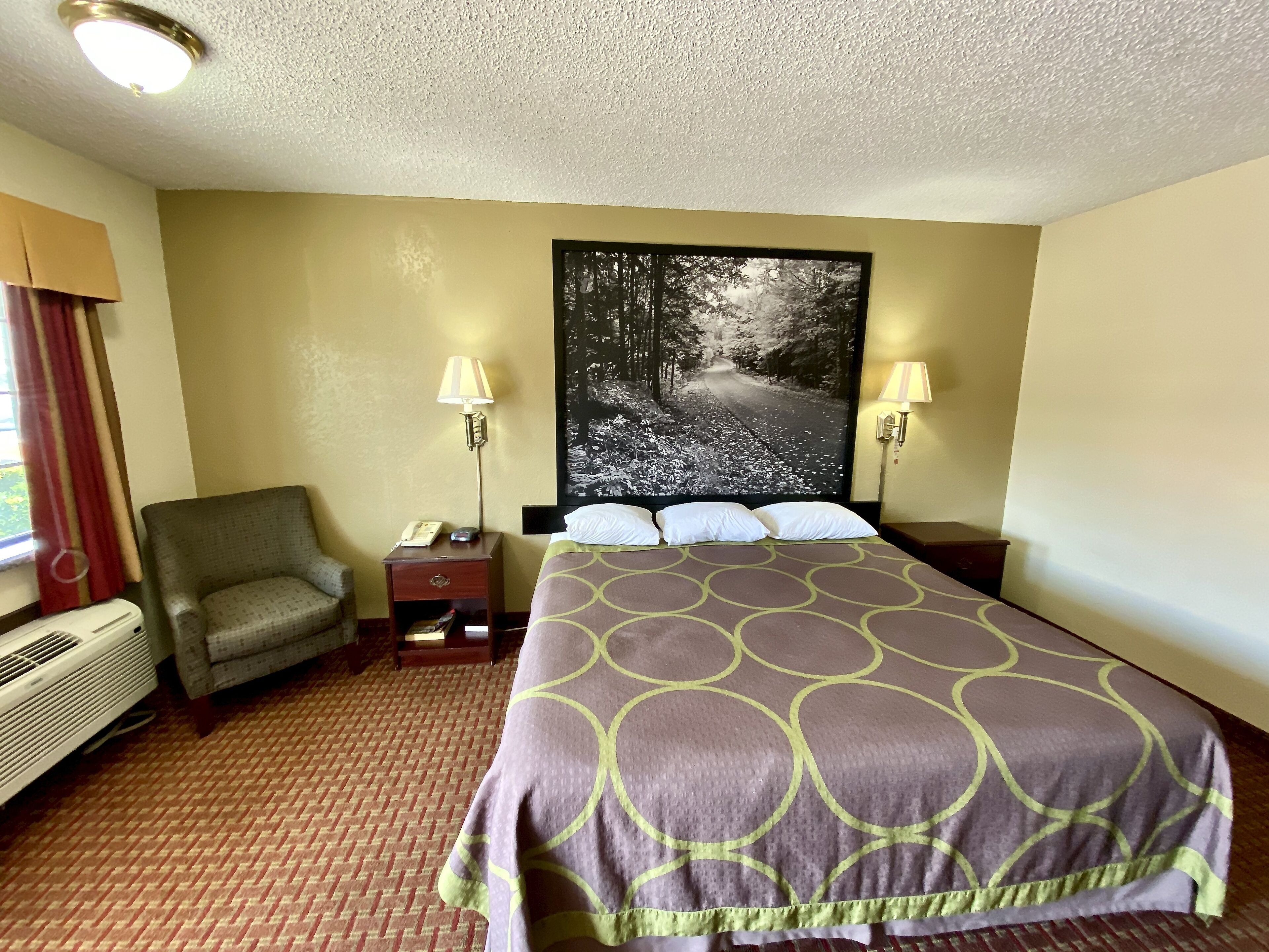 Habitación de hotel con cama, una silla, mesita de noche, lámpara, calefacción y ventana con cortinas.