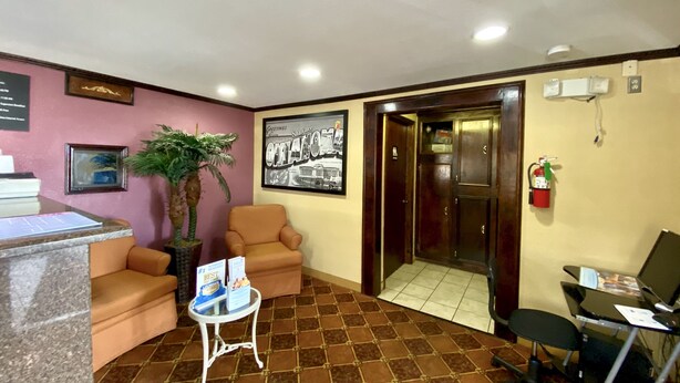 A reception area with a reception desk, two armchairs, a small table with brochures, a potted plant, a framed picture, a fire extinguisher, and a door leading to another room. - Super 8 by Wyndham Shawnee (Shawnee)