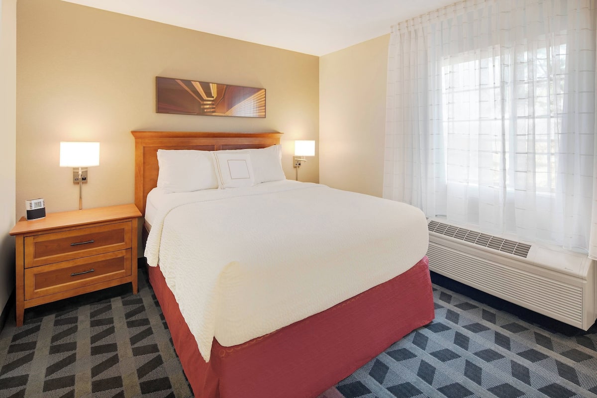 Une chambre d’hôtel avec un grand lit, deux tables de chevet avec des lampes, une œuvre d’art accrochée au mur, une fenêtre avec des rideaux légers et une moquette à motifs.