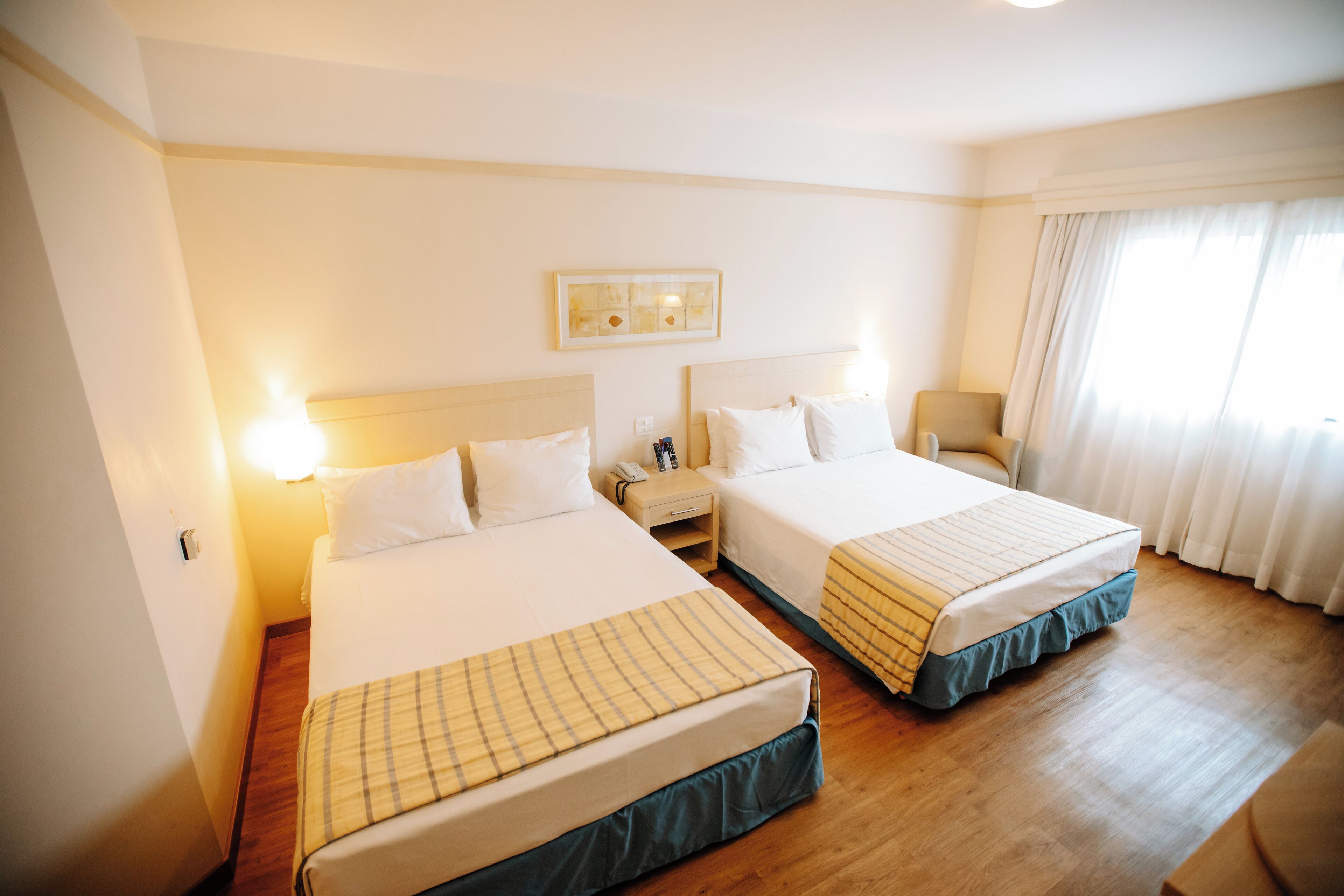 Quarto de hotel com duas camas, piso de madeira, mesa de cabeceira com abajur e janela com cortinas.