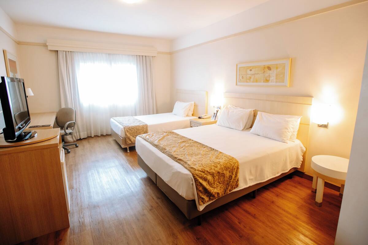 A hotel room with two beds, a desk with a computer, a chair, a TV, and a window with curtains.