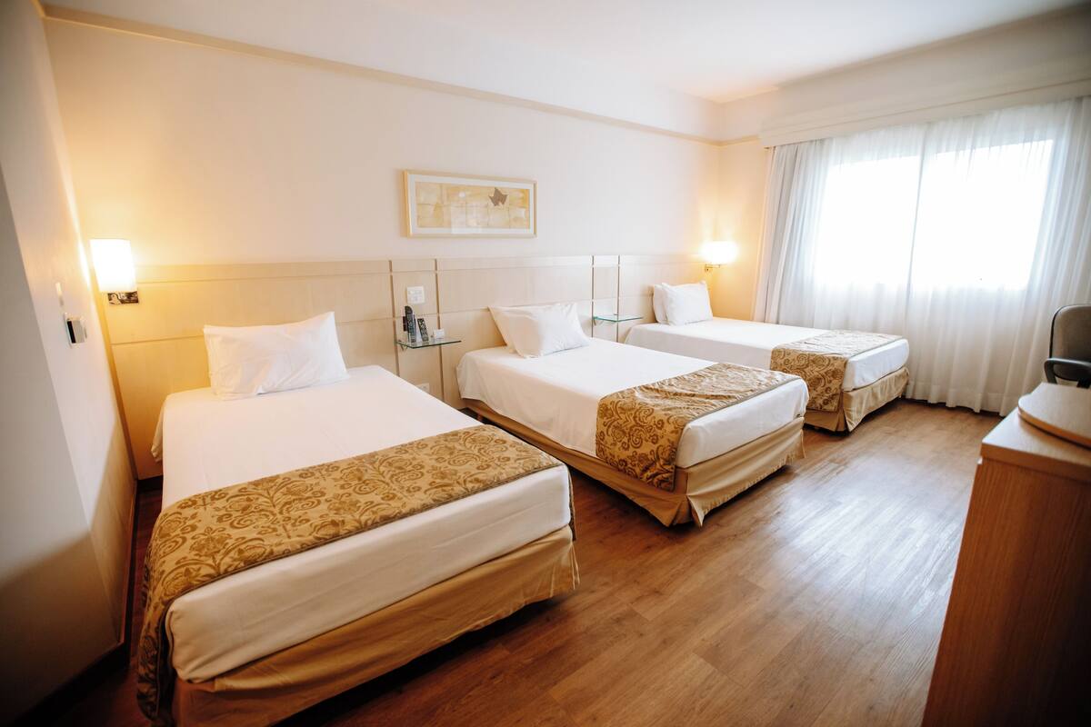 A hotel room with three single beds, wooden flooring, a bedside table with a lamp, and a large window with curtains.