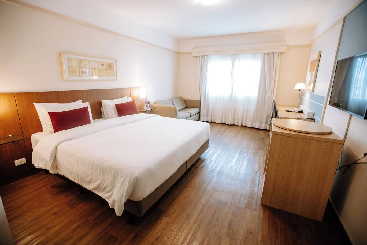 A hotel room with a large bed, a desk with a chair, a TV, and a window with curtains.