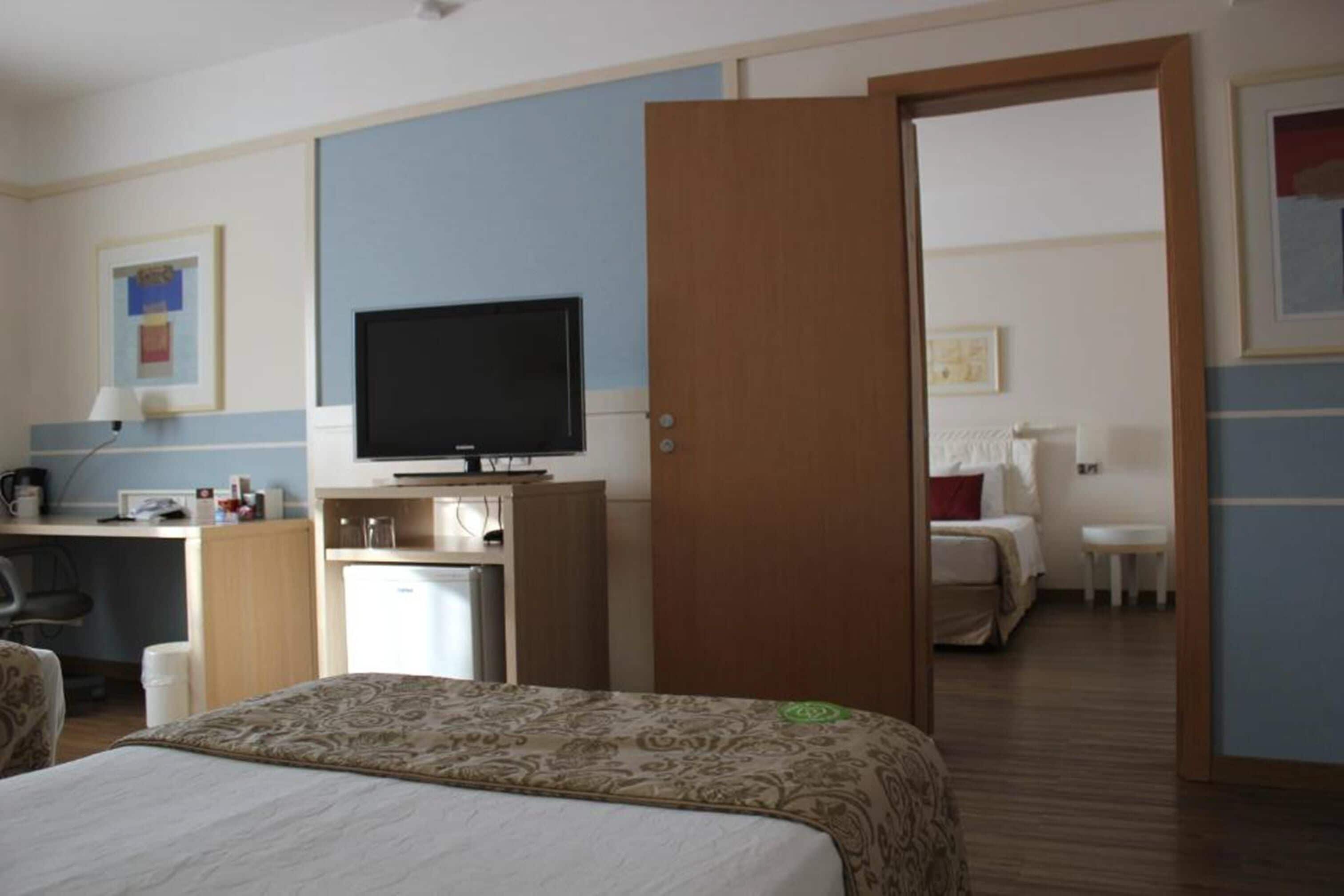 A hotel room with a bed, a desk with a television, a small refrigerator, and a door leading to another room.