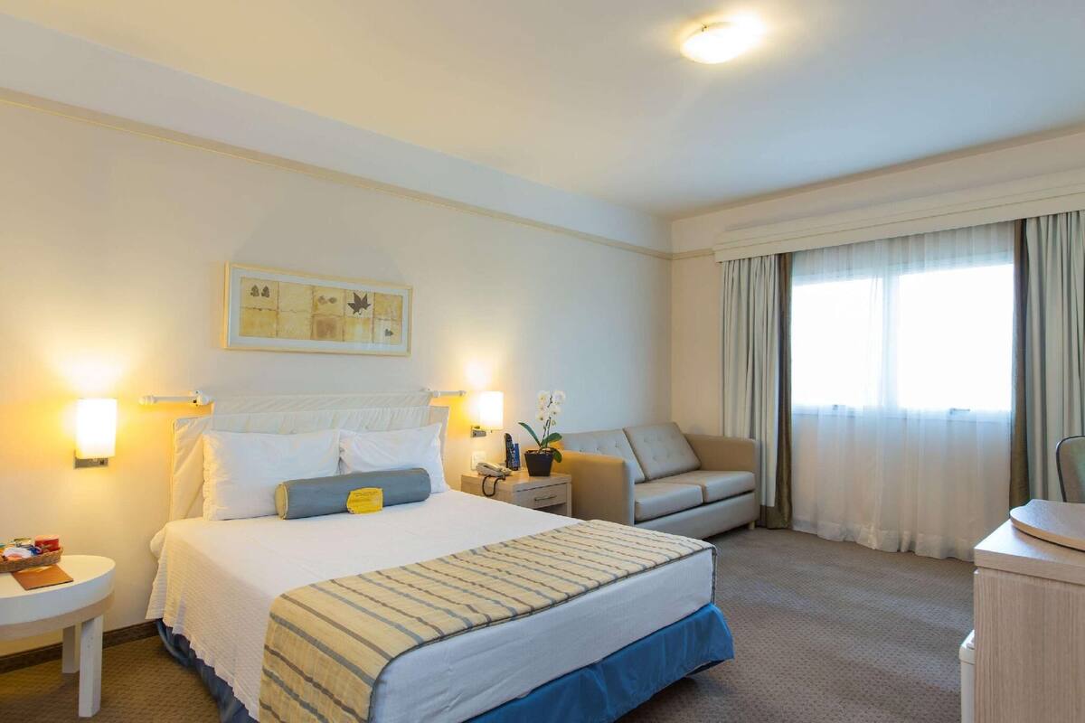 Una habitación de hotel con cama, dos mesitas de noche, sofá, escritorio y ventana con cortinas.