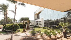 Un bâtiment moderne à la façade vitrée, entouré d’un jardin bien entretenu où l’on trouve des palmiers et diverses plantes.