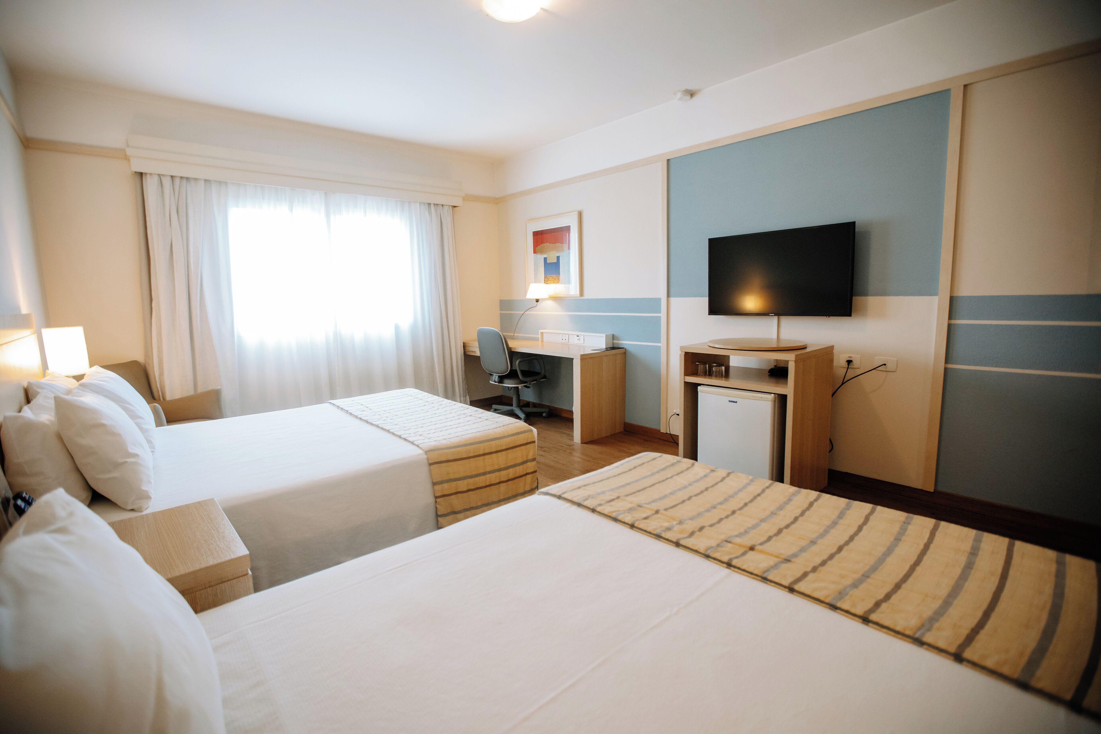 Quarto de hotel com duas camas, uma escrivaninha, televisão e uma pequena cozinha.
