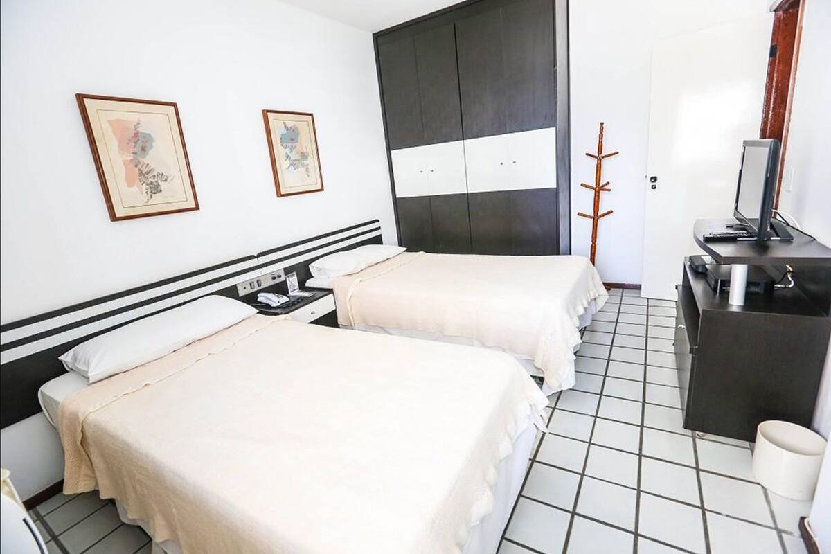 A hotel room with two beds, a desk with a computer, a wardrobe, and two framed pictures on the wall.